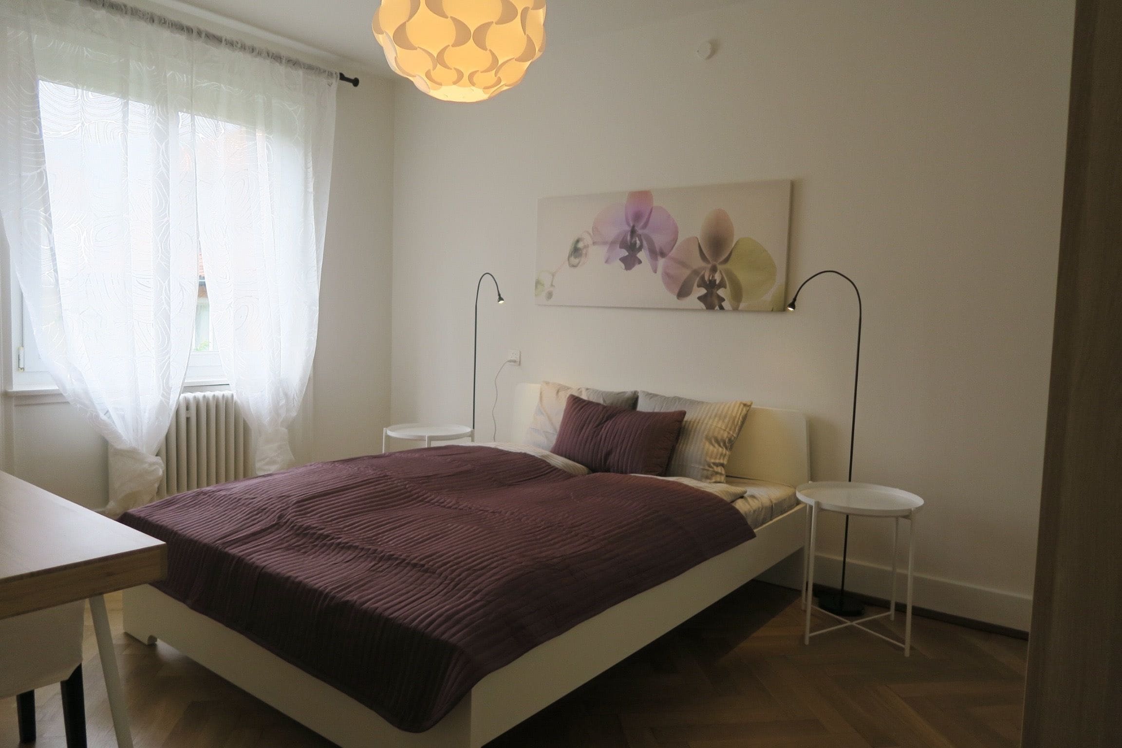 Une chambre à coucher comprenant un lit, des tables de chevet, un radiateur, une fenêtre avec des rideaux légers et un tableau représentant des fleurs accroché au mur.