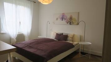Une chambre à coucher comprenant un lit, des tables de chevet, un radiateur, une fenêtre avec des rideaux légers et un tableau représentant des fleurs accroché au mur.