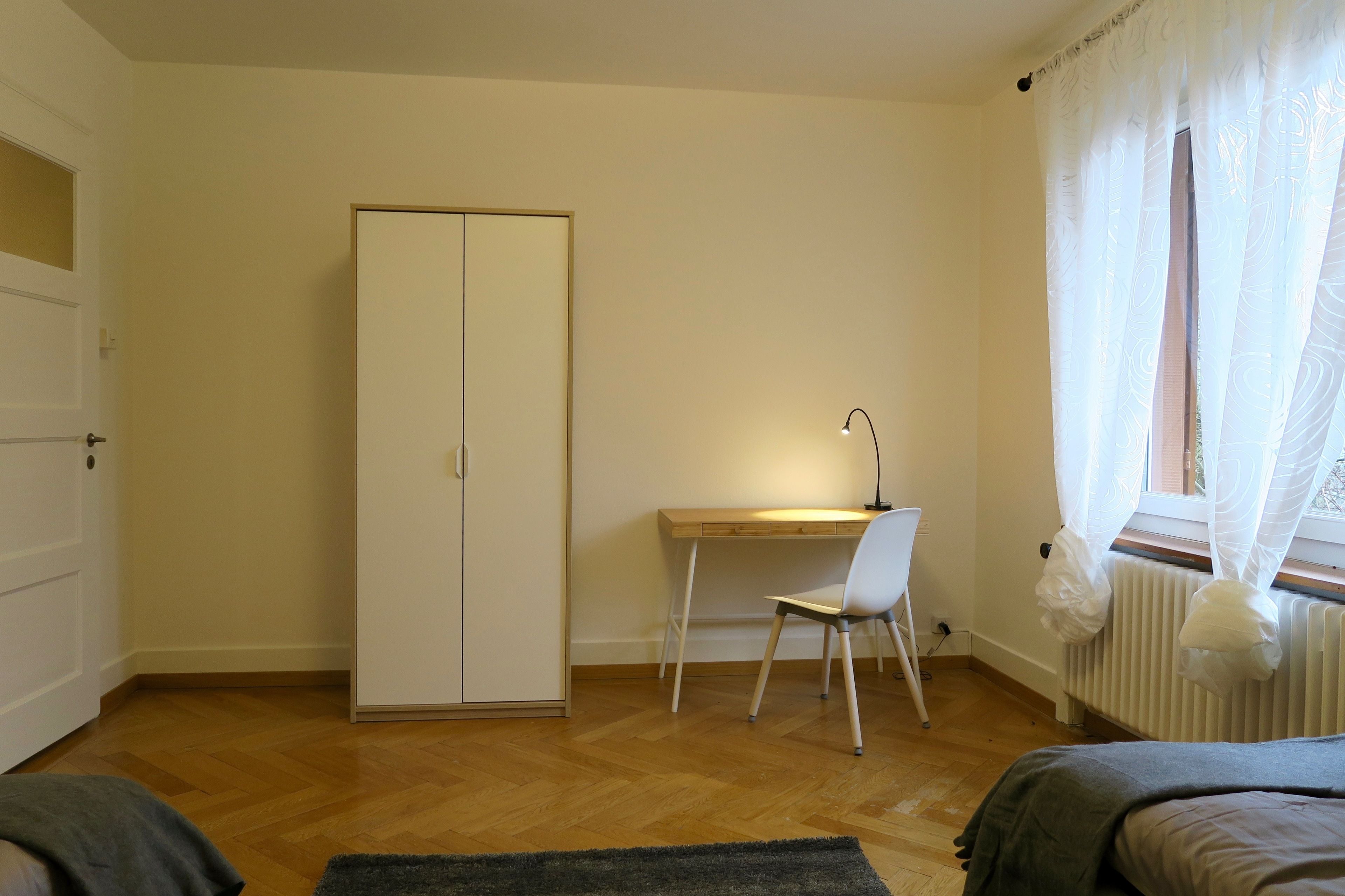 A bedroom with a wooden floor, a bed, a desk with a chair, a wardrobe, and a window with curtains.