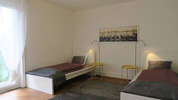 Un dormitorio con dos camas individuales, una mesita, una silla y una ventana con cortinas.