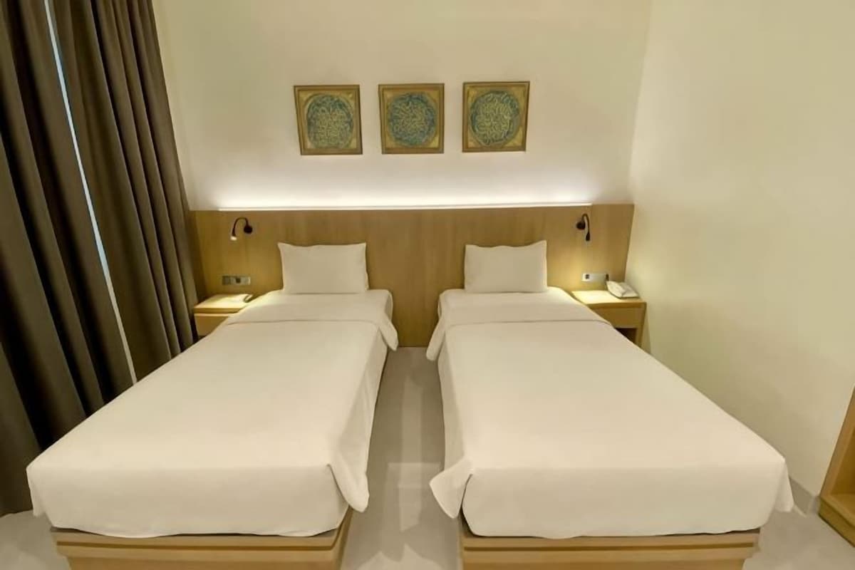 A hotel room with two single beds, a nightstand with a lamp, and a wall-mounted artwork.