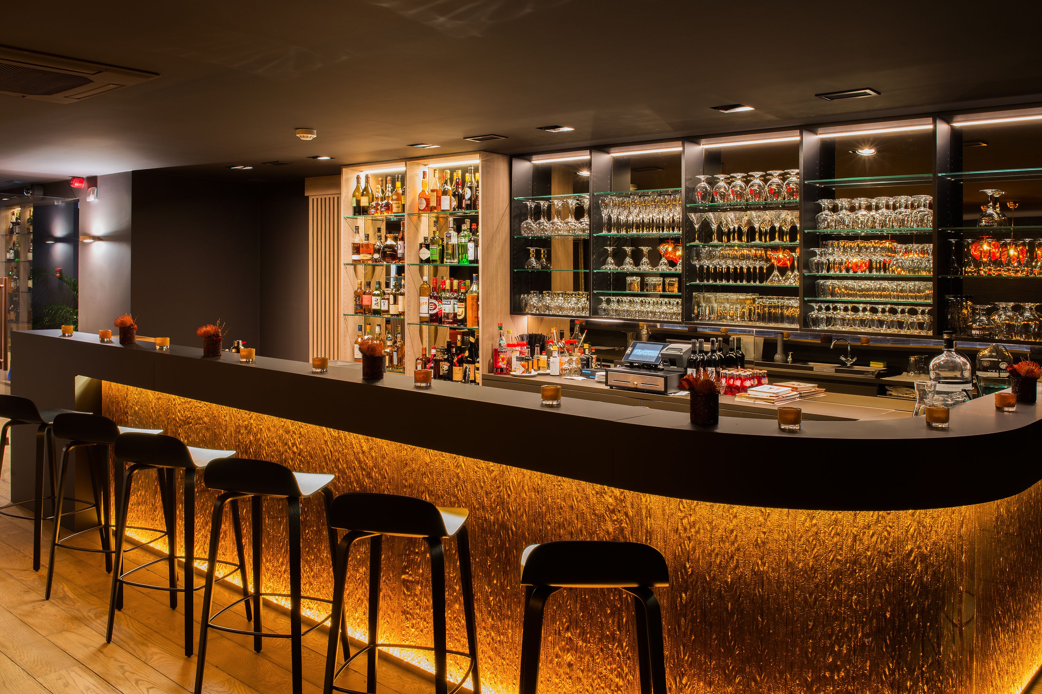 Un espace bar doté d’un comptoir incurvé, de tabourets de bar et d’étagères remplies de bouteilles et de verres.