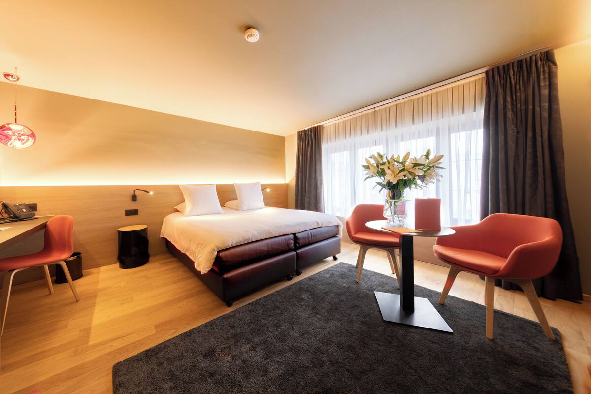 A modern hotel room with a bed, desk, chairs, and a small table with a vase of flowers.