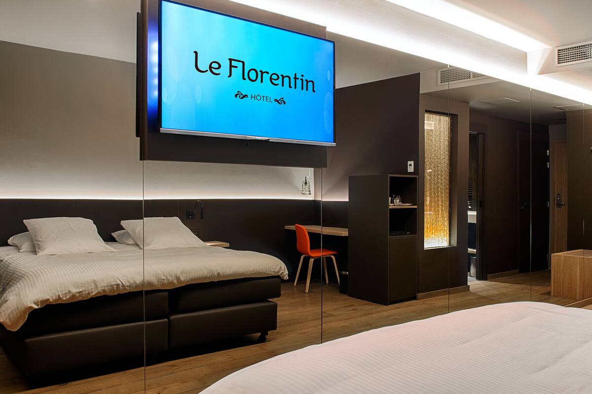 A modern hotel room with a large bed, a desk, and a television displaying the hotel name.