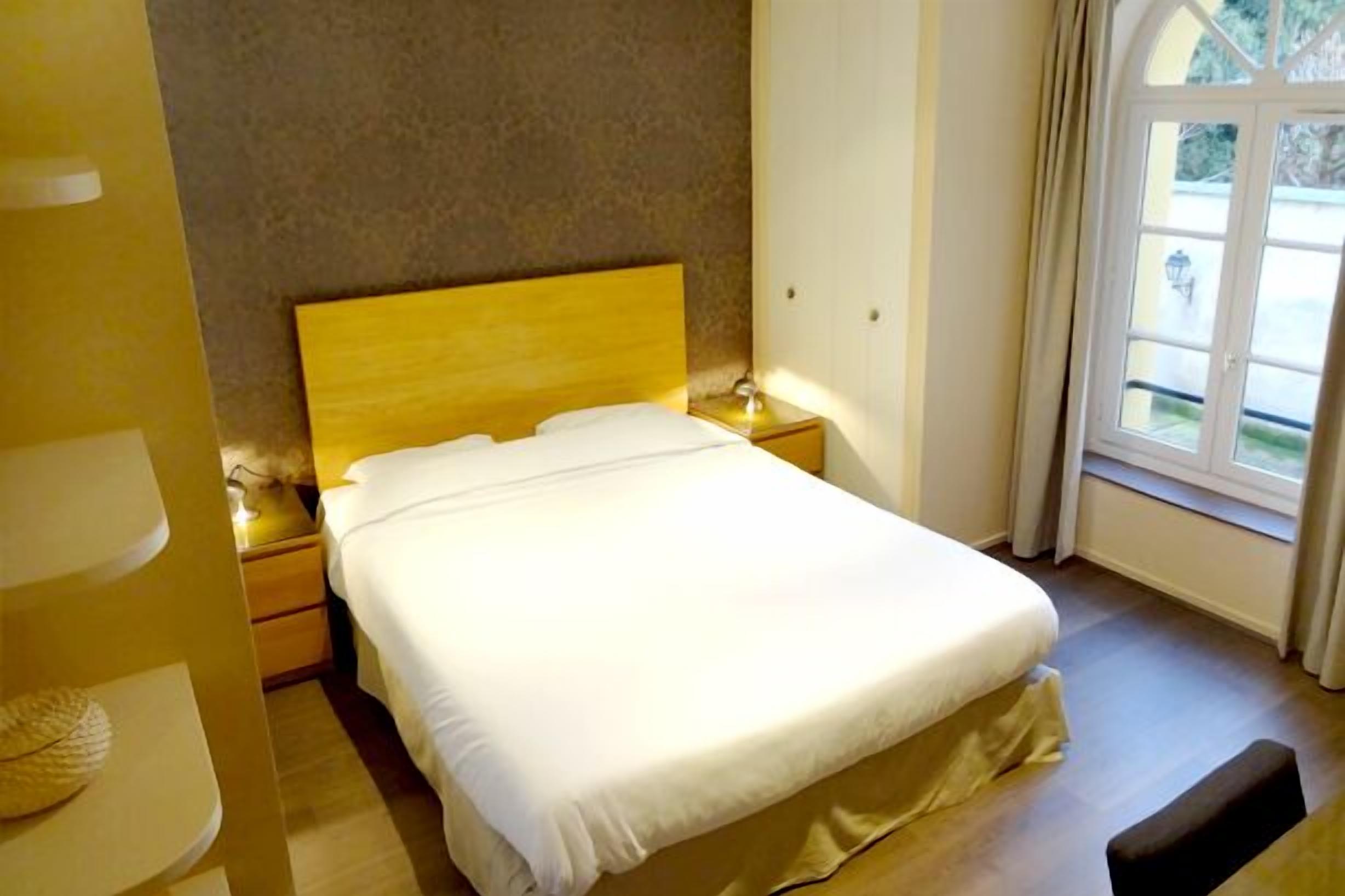 Un letto rifatto con cura, con una testiera in legno, un comodino con una lampada e una finestra con tende.