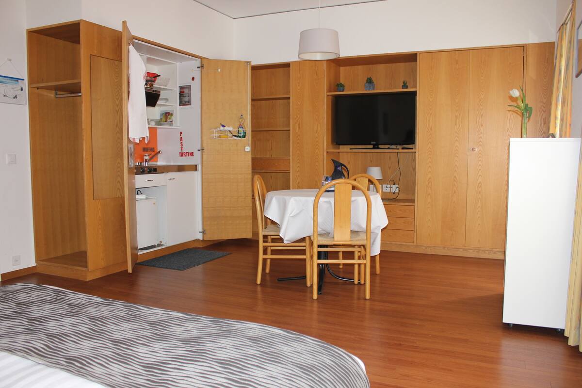 A compact room with a bed, a dining table with chairs, a kitchenette, and a television.