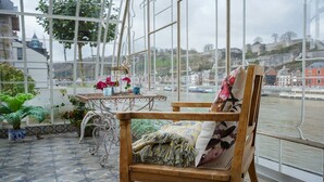 Una silla de madera con un cojín floral, una mesita con un florero y vistas a un río con edificios al fondo.