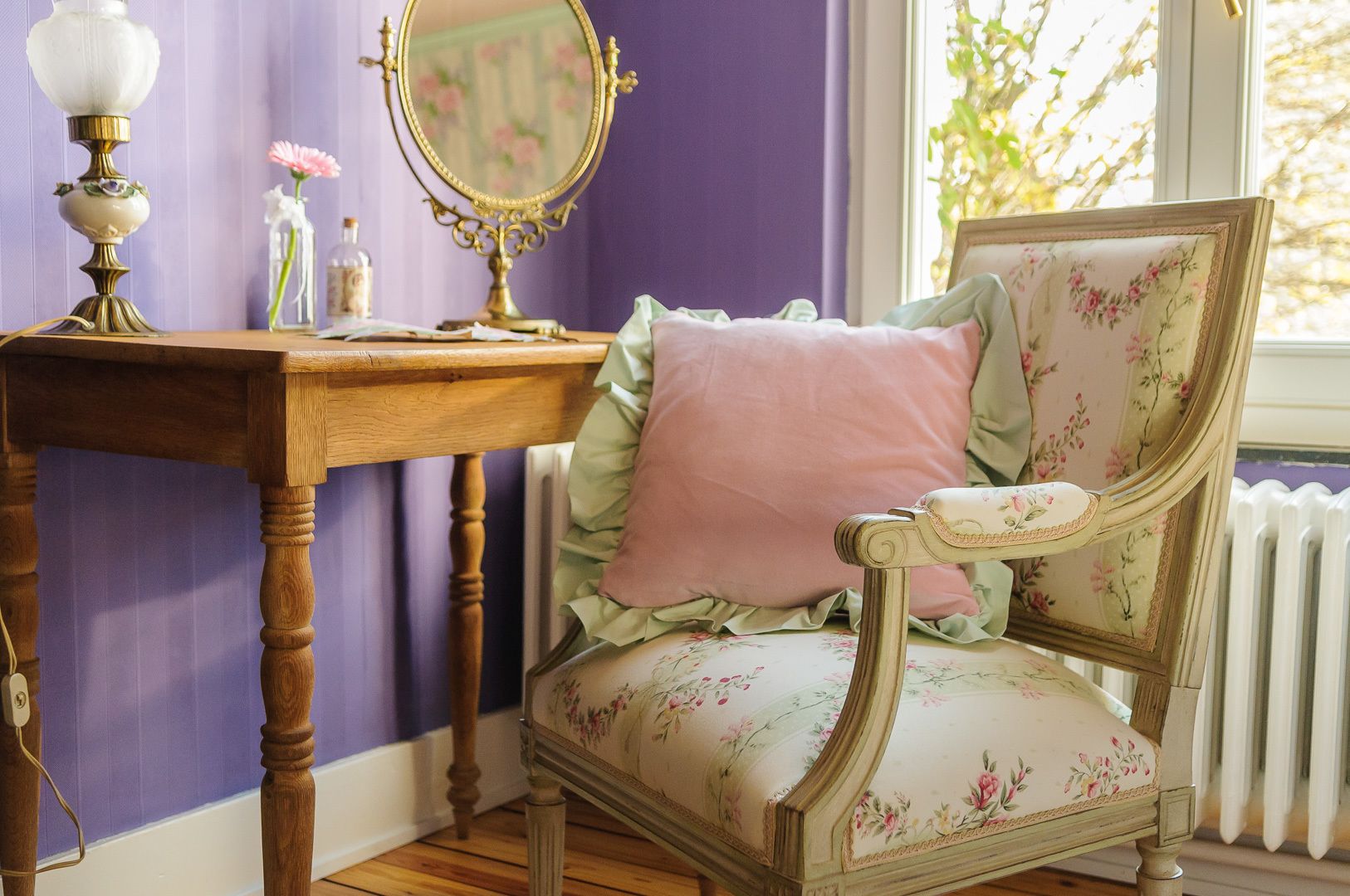 Une chaise de style vintage, recouverte d’un tissu à motifs floraux, une table en bois avec une lampe et un miroir orné d’un cadre floral.