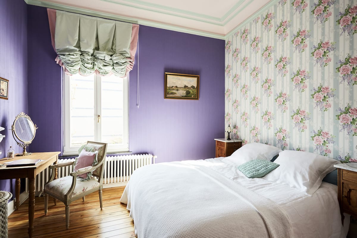 A bedroom with a bed, a chair, a dresser, a window with curtains, and a radiator.