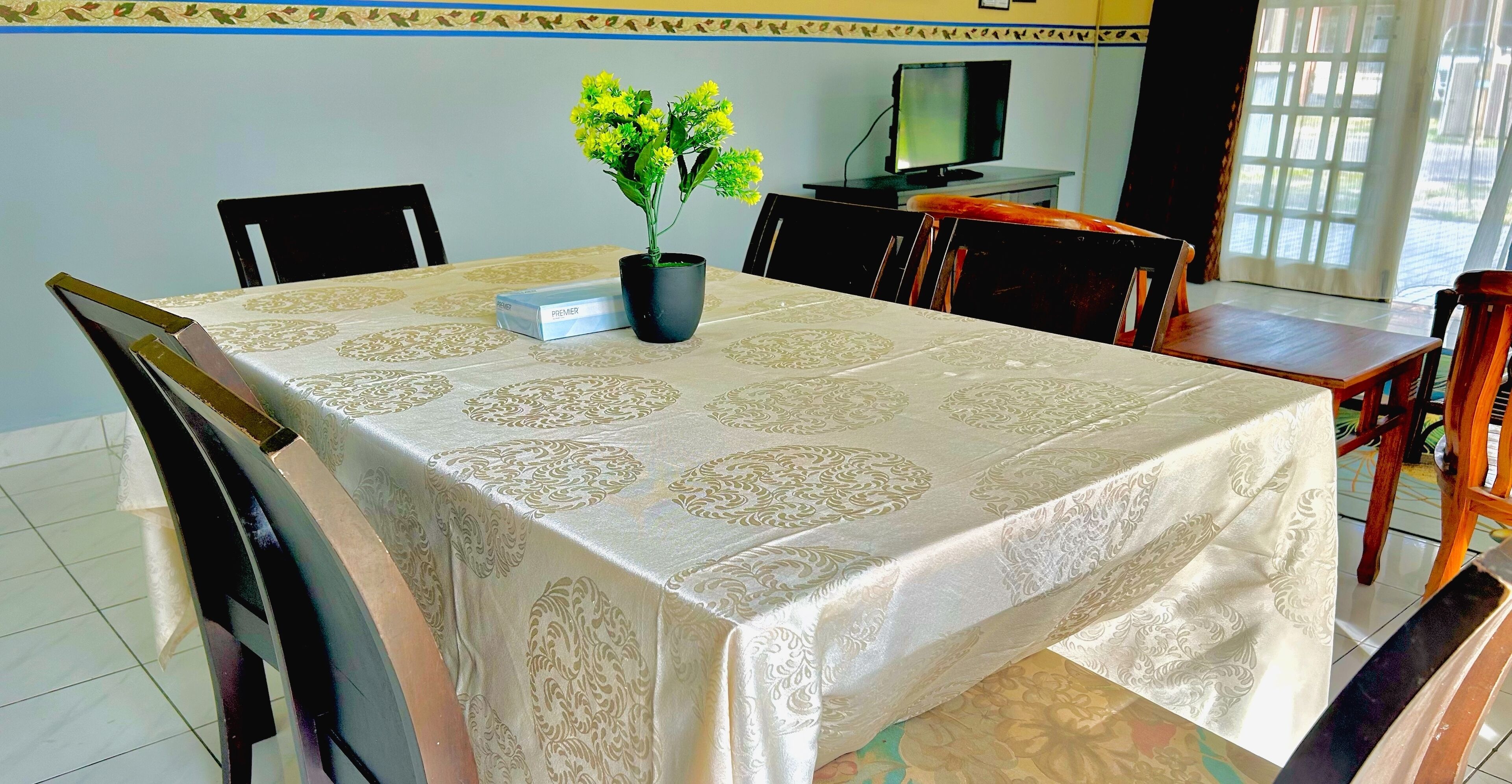 Un comedor con una mesa cubierta por un mantel estampado, una planta en maceta y un televisor al fondo.