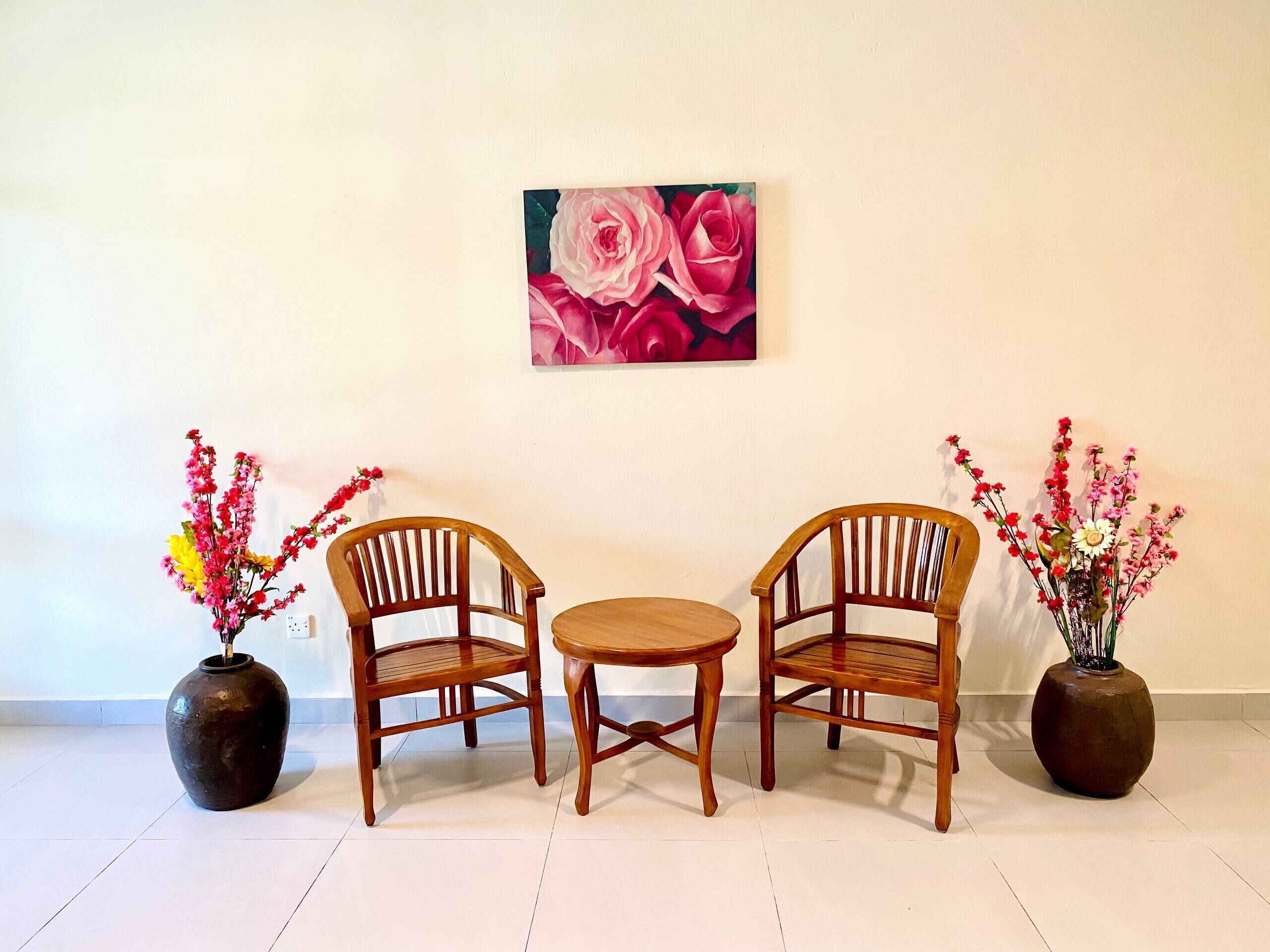 Een kamer met twee houten stoelen en een kleine ronde tafel, een schilderij van roze rozen aan de muur, en twee vazen met bloemen op de vloer.