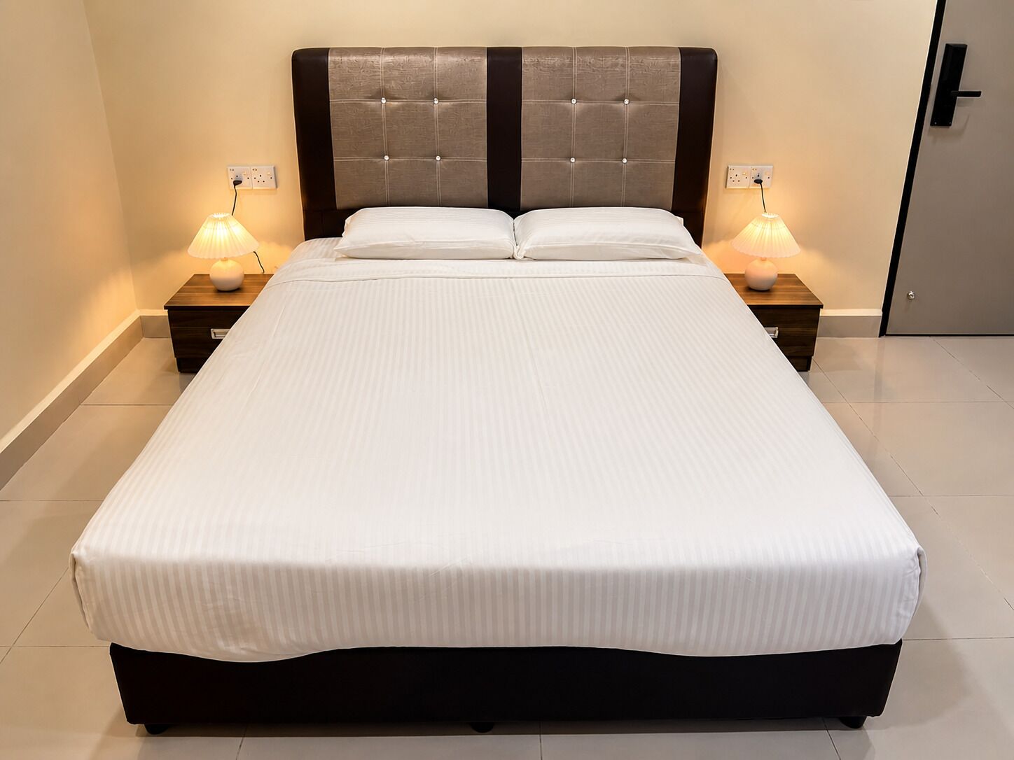 Una cama grande con cabecera capitonada, dos mesitas de noche con lámparas, y una puerta a la derecha.