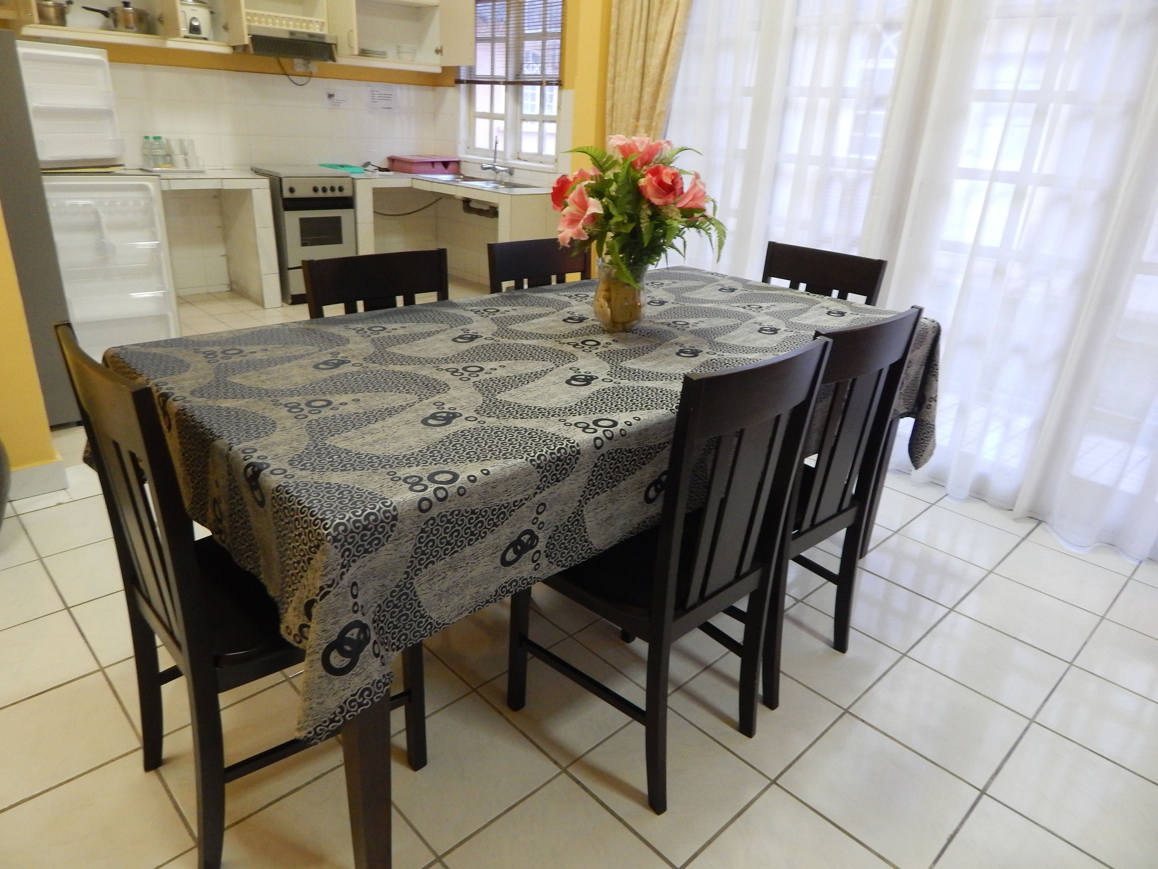 A dining table with a patterned tablecloth, surrounded by chairs, in a room with a kitchen in the background.