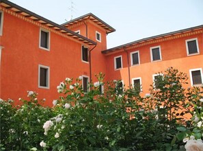 A building with a red facade and multiple windows, surrounded by lush greenery and white flowers. - Villa Chiopris (Chiopris-Viscone)