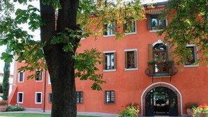 Ein rotes Gebäude mit einem mittleren Eingang, zahlreichen Fenstern und einem großen Baum im Vordergrund.