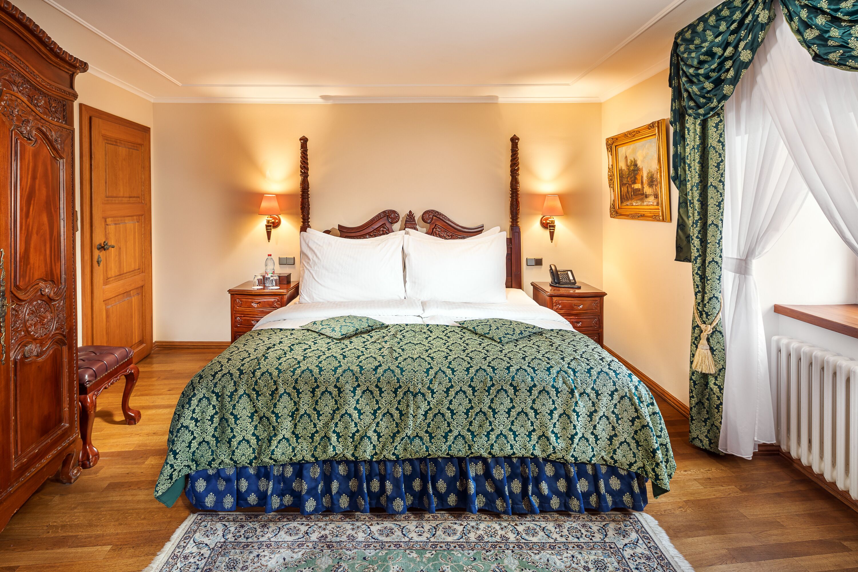 Ein Schlafzimmer mit einem großen Bett, einem Holz-Kopfteil, zwei Nachttischen mit Lampen, einem Beistelltisch, einer Bank, einem Bild an der Wand und einem Fenster mit Vorhängen.