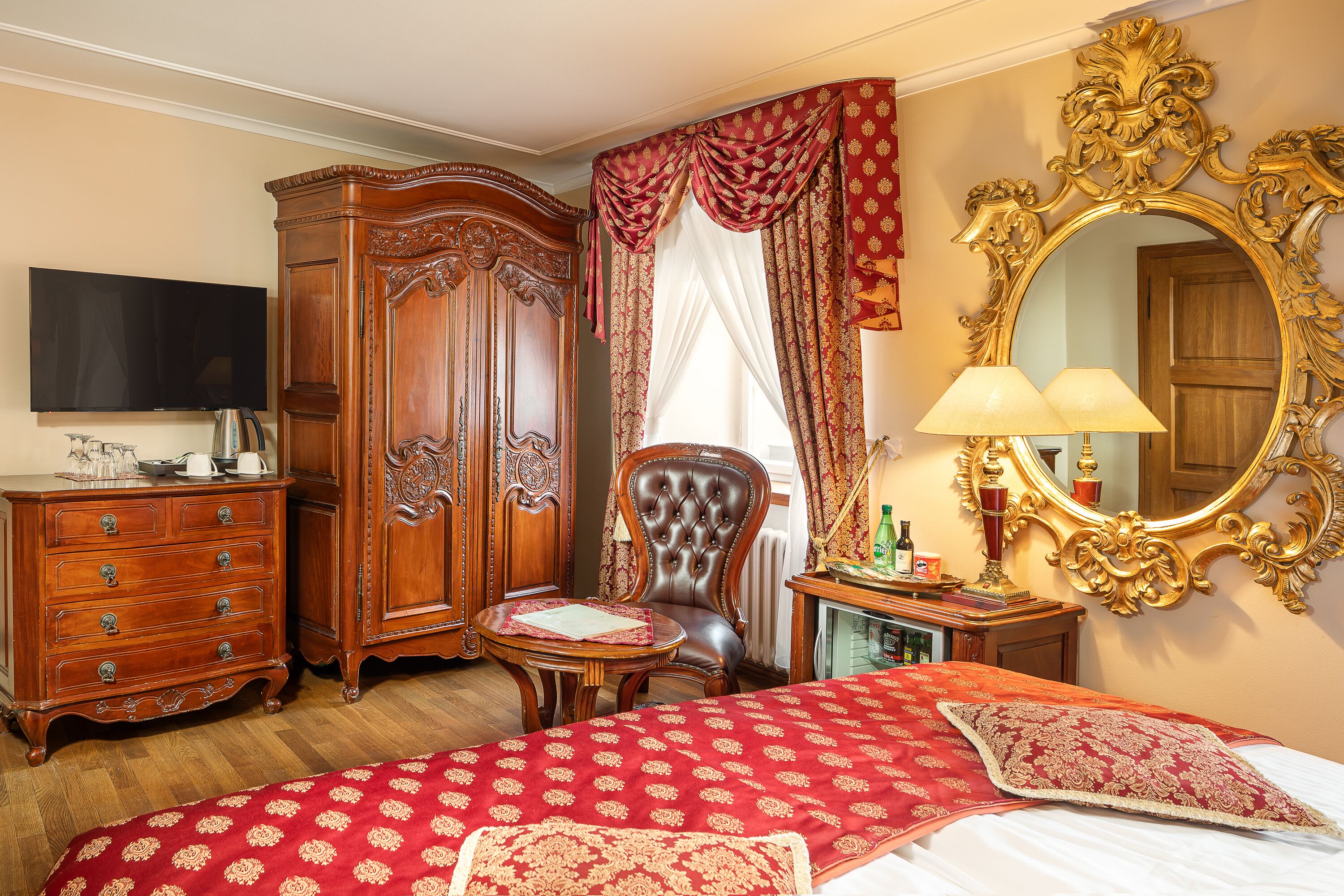 A bedroom with a large bed, a wooden wardrobe, a chair, a table, a mirror, and a television.