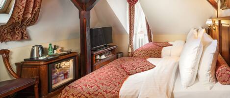 A bedroom with a sloped ceiling, a bed with white and red bedding, a wooden dresser, a television, and a window with curtains.