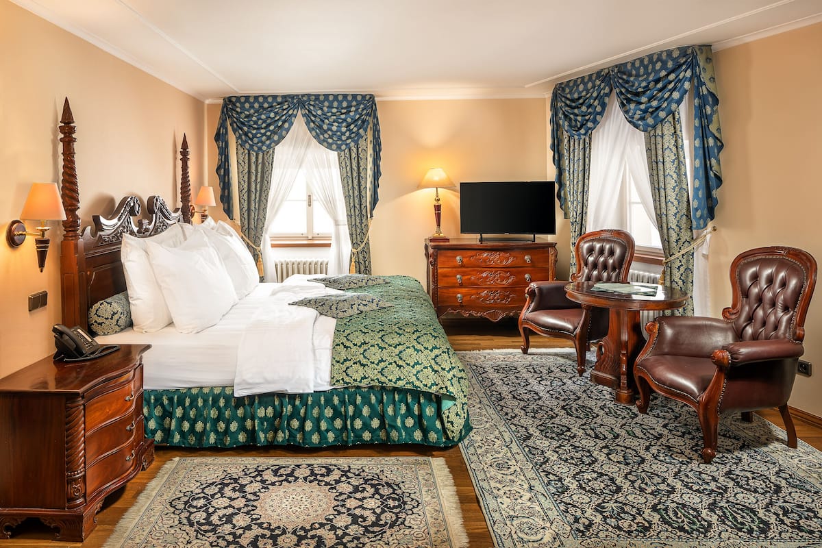 Une chambre d’hôtel avec un grand lit, une commode en bois, un téléviseur, deux chaises et un tapis à motifs.