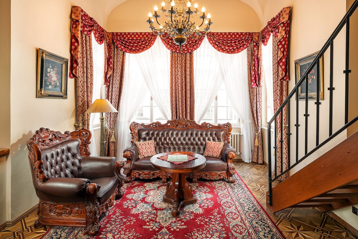 Une vaste pièce comprenant un grand canapé richement décoré, un fauteuil assorti et une table basse ronde en bois. Un lustre est suspendu au plafond, et un escalier est visible sur la droite.