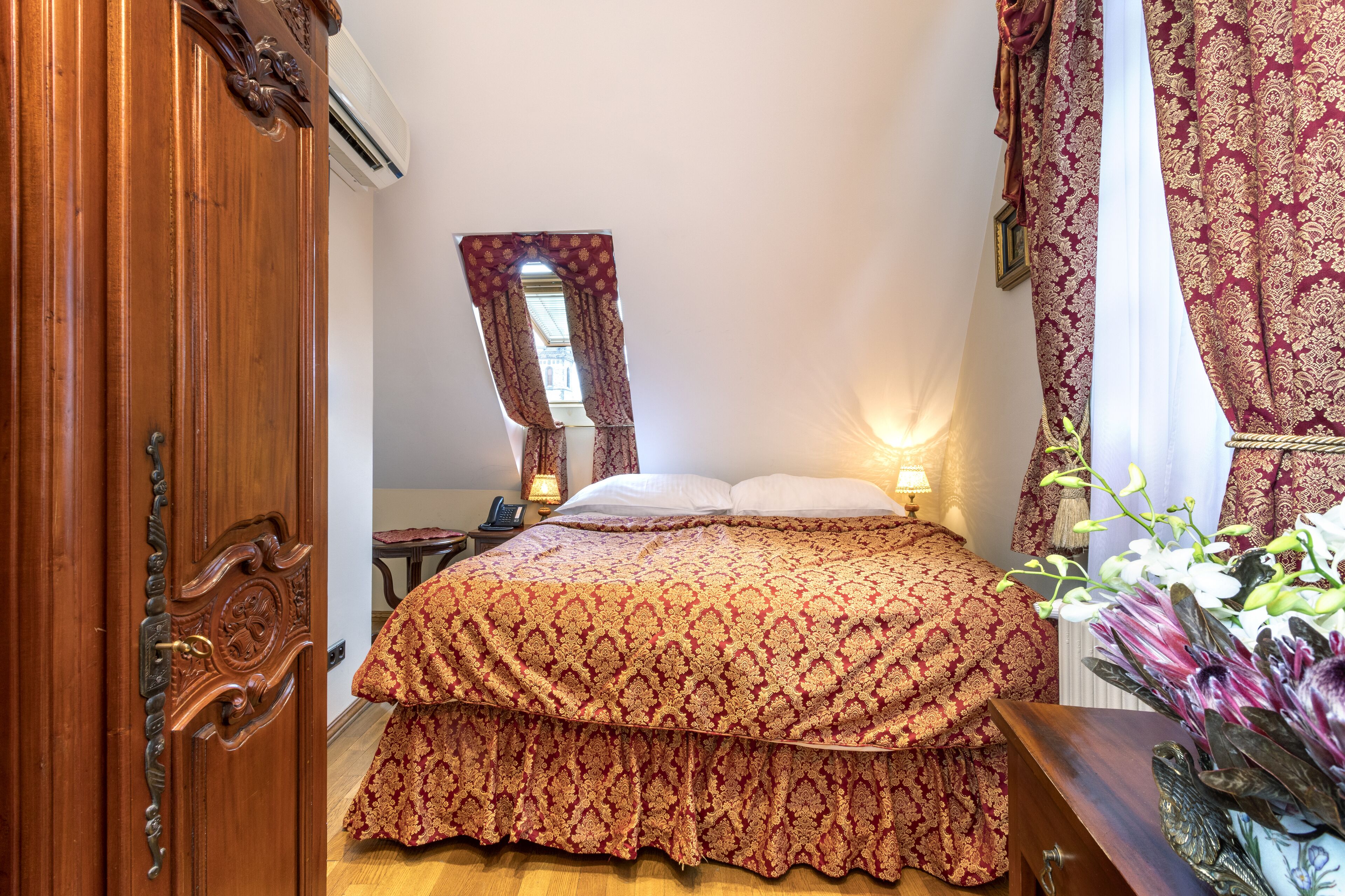 A bedroom with a wooden wardrobe, a bed with a patterned bedspread, a nightstand with a lamp, and a window with curtains.