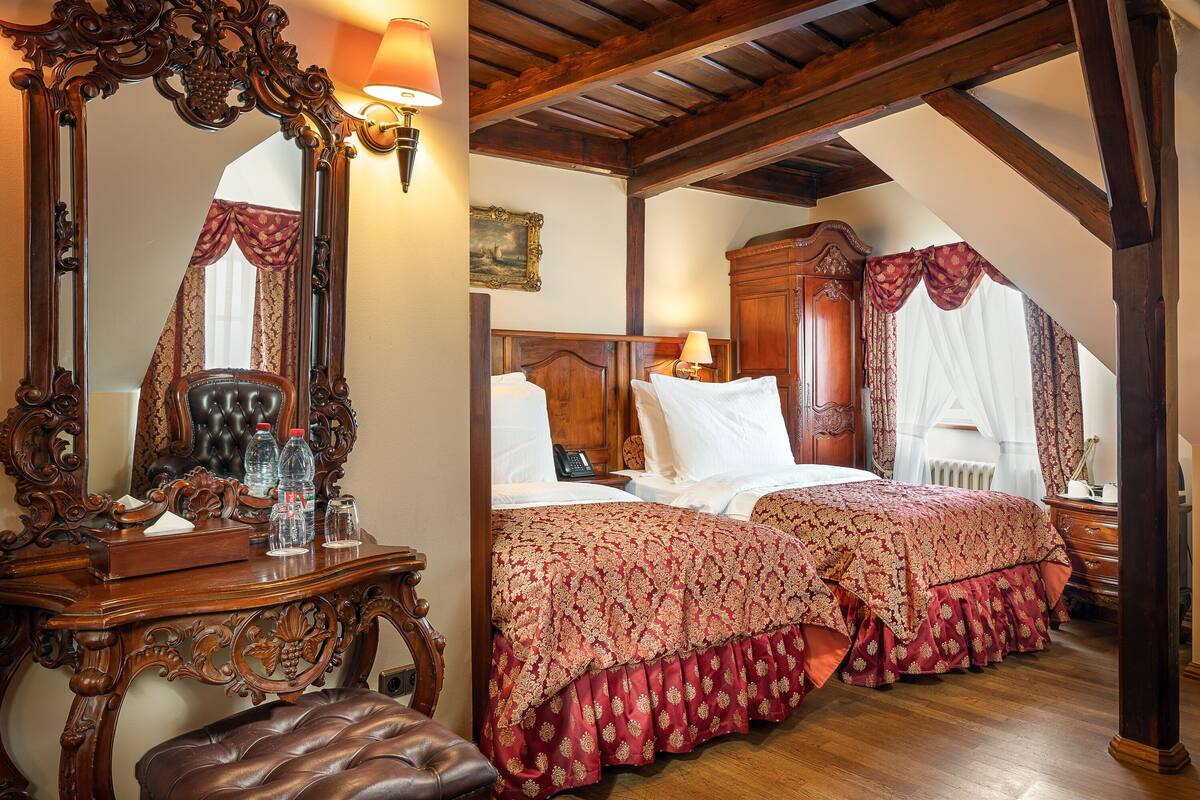 Une chambre à coucher avec un grand miroir décoratif, une tête de lit en bois, deux lits avec une literie à motifs rouges, une commode en bois et un banc.