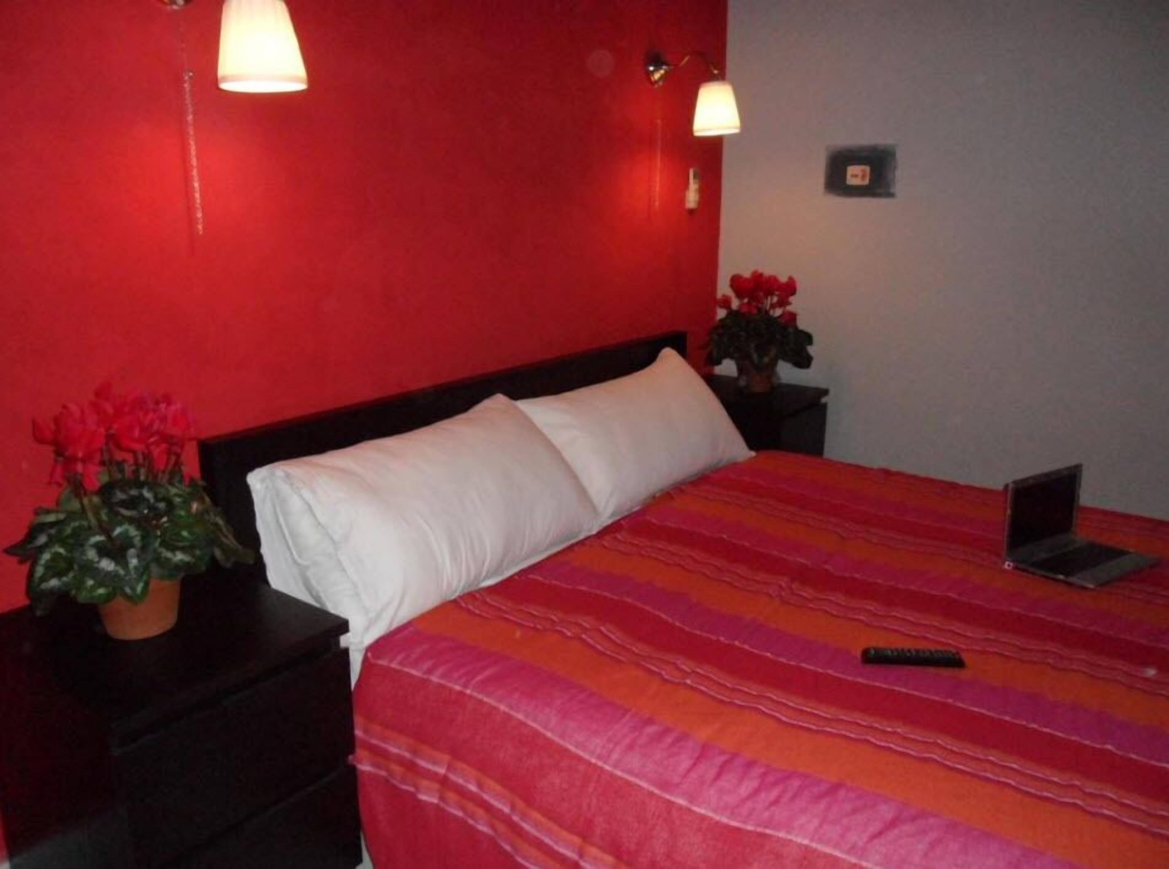 Une chambre d’hôtel avec un mur rouge, un lit recouvert d’une couverture à rayures, un oreiller blanc, deux tables de chevet avec des lampes, un ordinateur portable sur l’une des tables et une plante en pot sur l’autre.