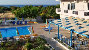 A pool area with lounge chairs and umbrellas, a covered seating area with tables and chairs, and a building with a tiled roof. - Greta Beach (Hersonissos)