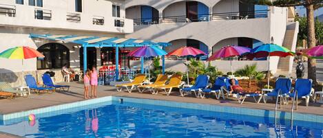 A pool area with multiple lounge chairs and umbrellas, a clear blue pool, and a building with balconies and air conditioning units.