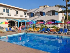 Un área de piscina con múltiples sillas de descanso y sombrillas, una piscina azul cristalina y un edificio con balcones y unidades de aire acondicionado.