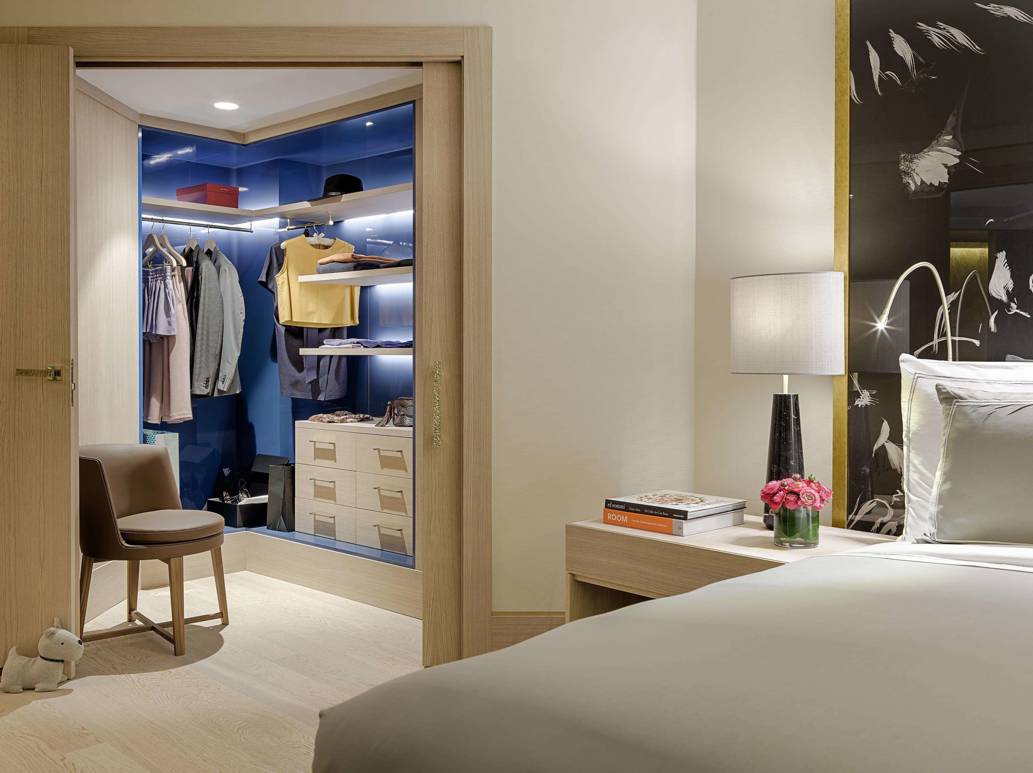 A hotel room with a bed, a desk with a lamp, a chair, a wardrobe with clothes, and a small table with books and flowers.