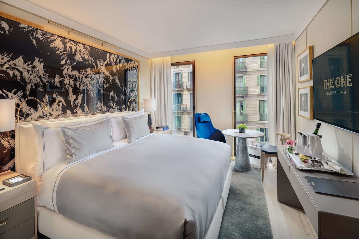 A modern hotel room with a large bed, a desk with a tray of breakfast items, a blue chair, and a view of the city.