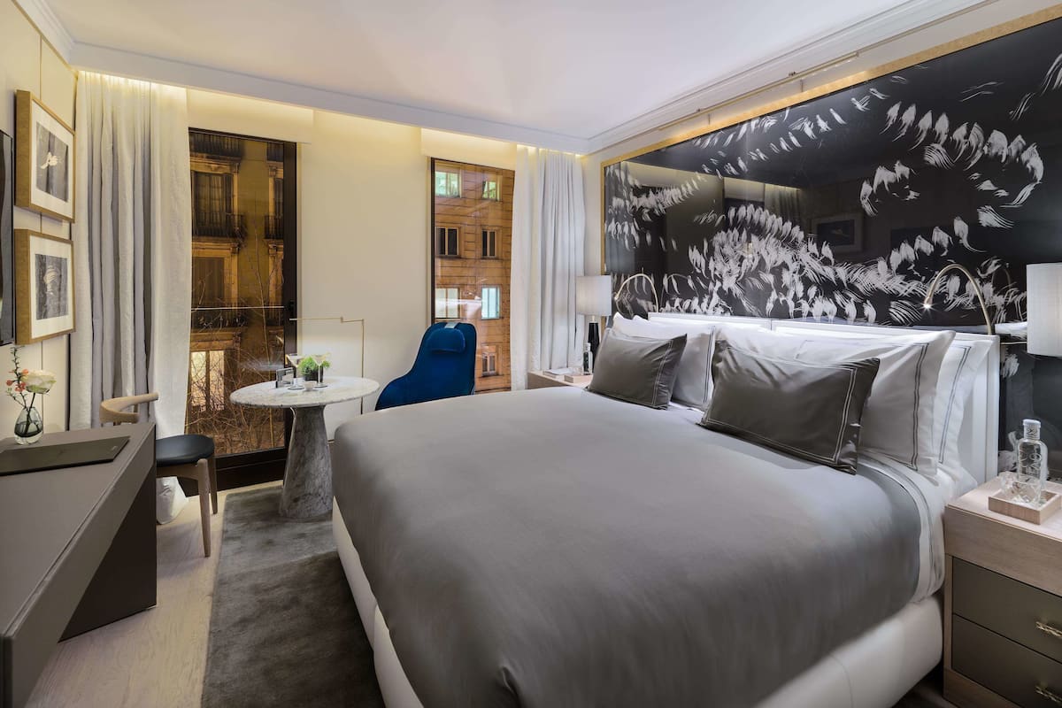 A modern hotel room with a large bed, a desk, a chair, and a small table.