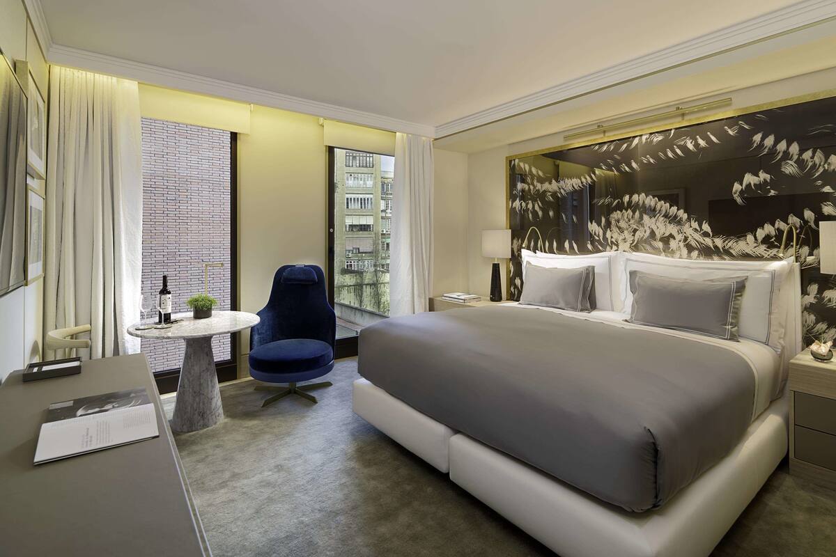 A modern hotel room with a large bed, a desk with a chair, a round table, and a view of the city through a large window.