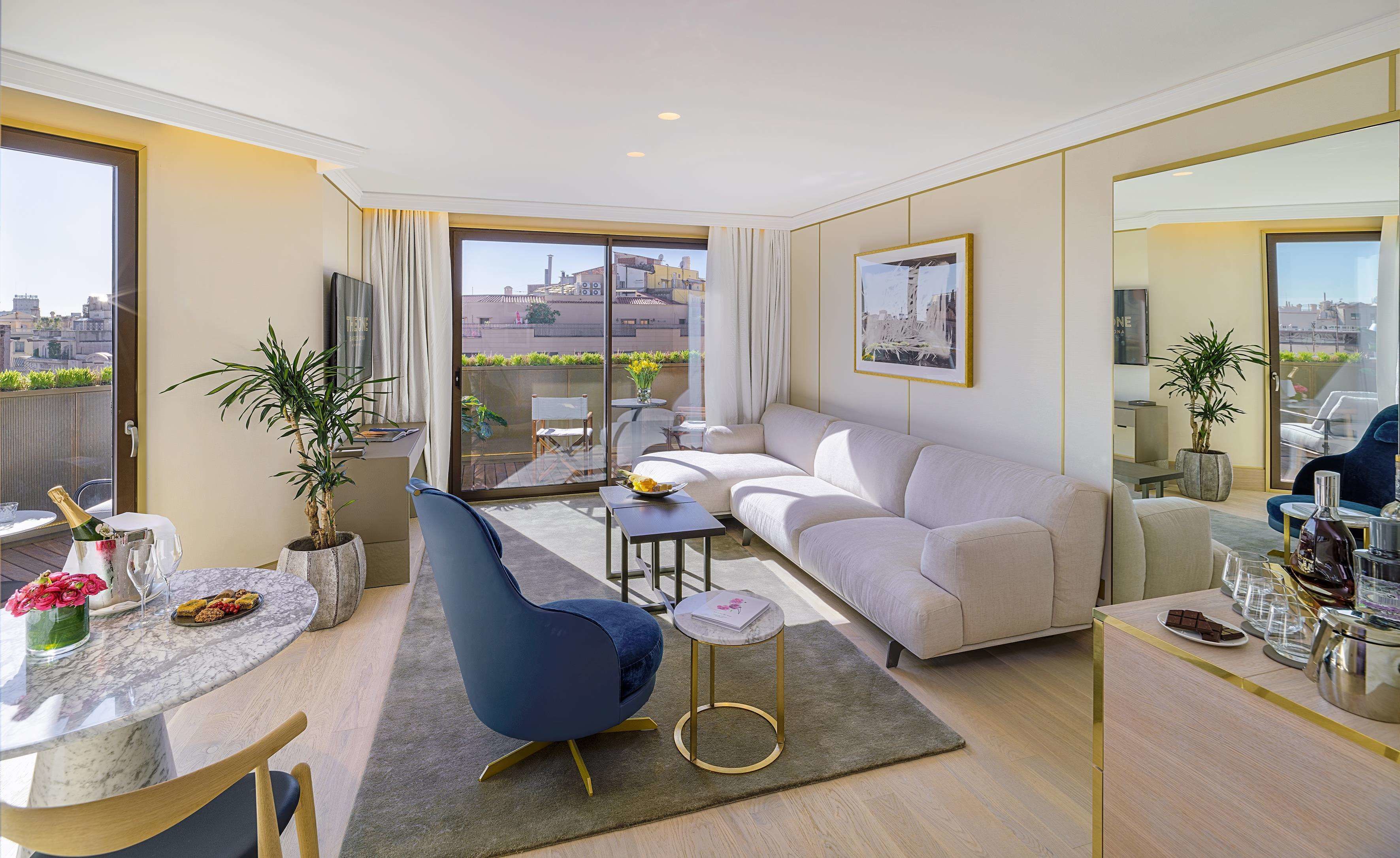 A modern living room with a sofa, a blue chair, a small table with a bottle and glasses, and a balcony with a view of buildings.