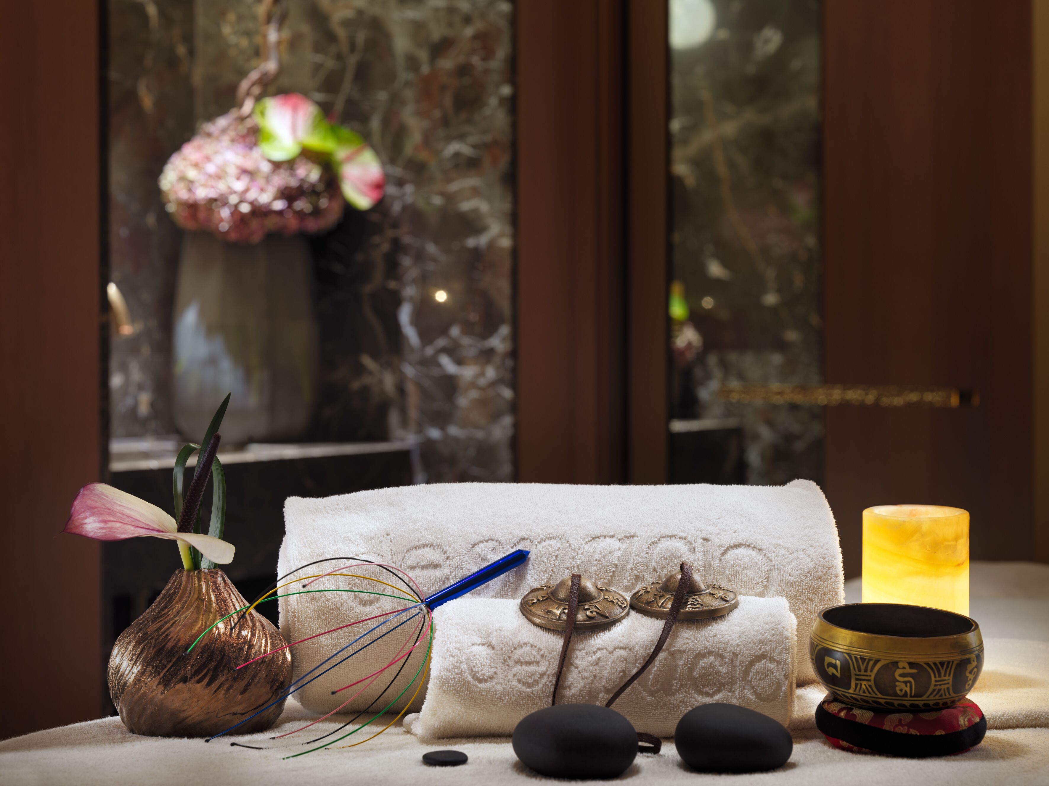 Un espacio de relajación con una toalla, un cuenco de meditación, una vela, un cuenco tibetano, un objeto con plumas y un jarrón con una flor.