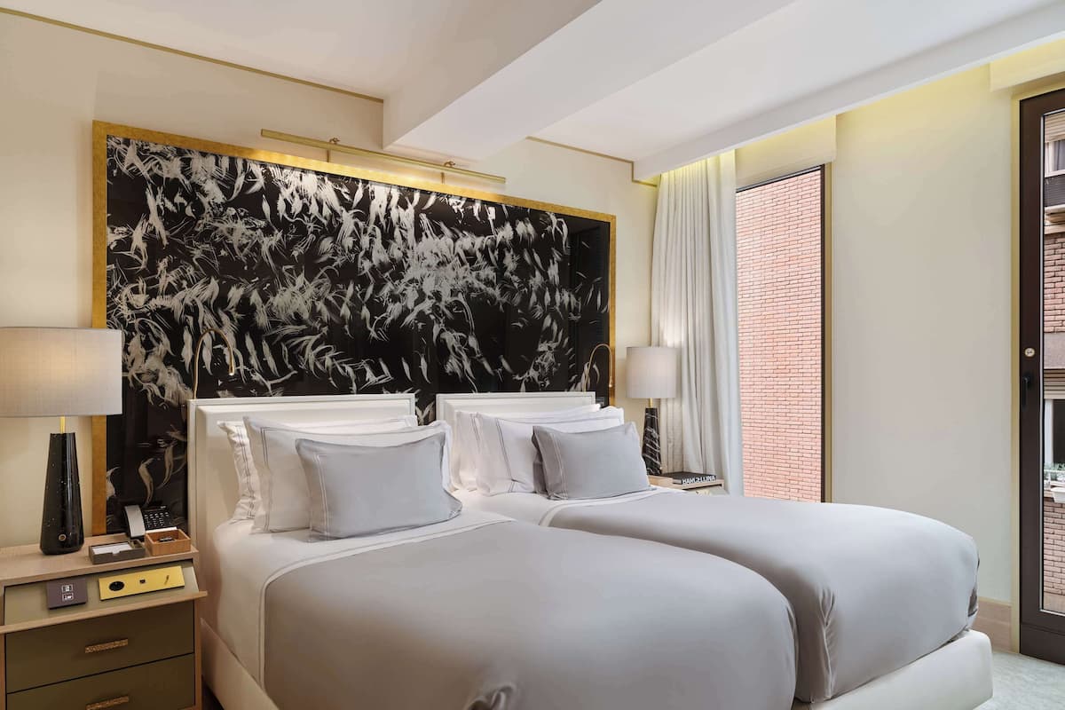 A hotel room with two beds, a large abstract painting on the wall, a nightstand with a lamp, and a view of a brick wall through a door.