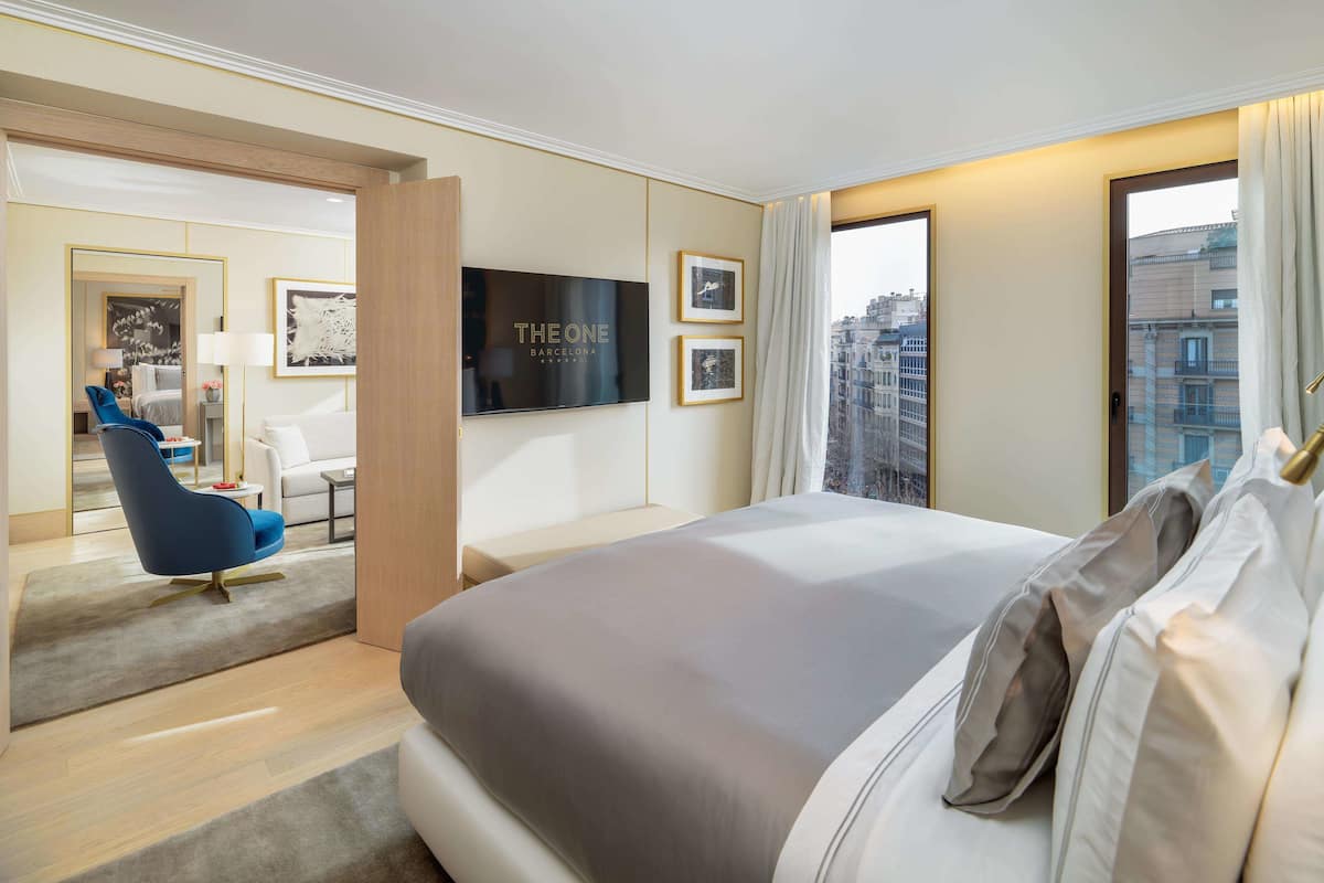 A modern hotel room with a large bed, a TV mounted on the wall, a seating area with a blue chair, and a view of the city through the window.
