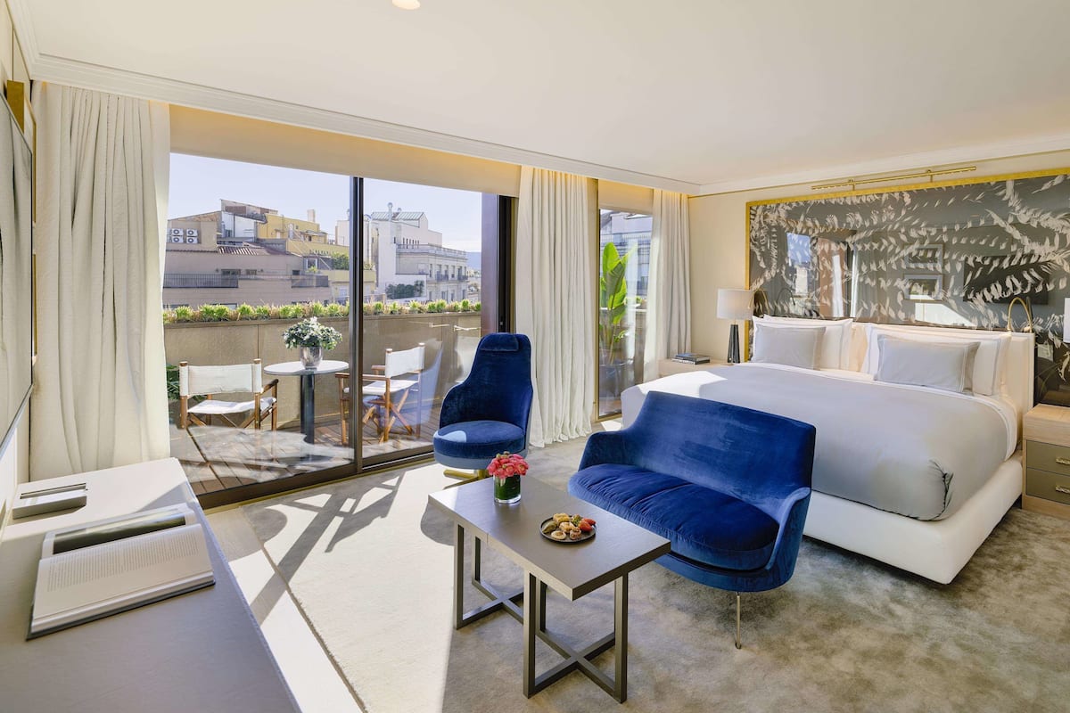 A modern hotel room with a large bed, a desk, two chairs, and a view of the city through large windows.