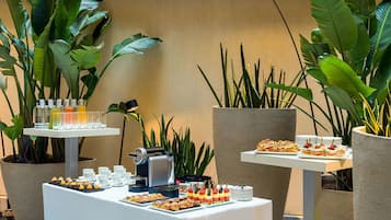 A buffet setup with various food items and beverages, surrounded by large potted plants.