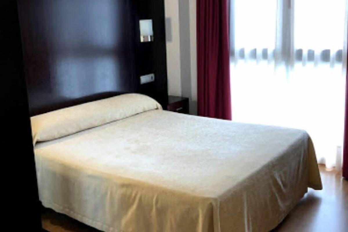 A neatly made bed with a headboard, a bedside table, and a window with curtains.