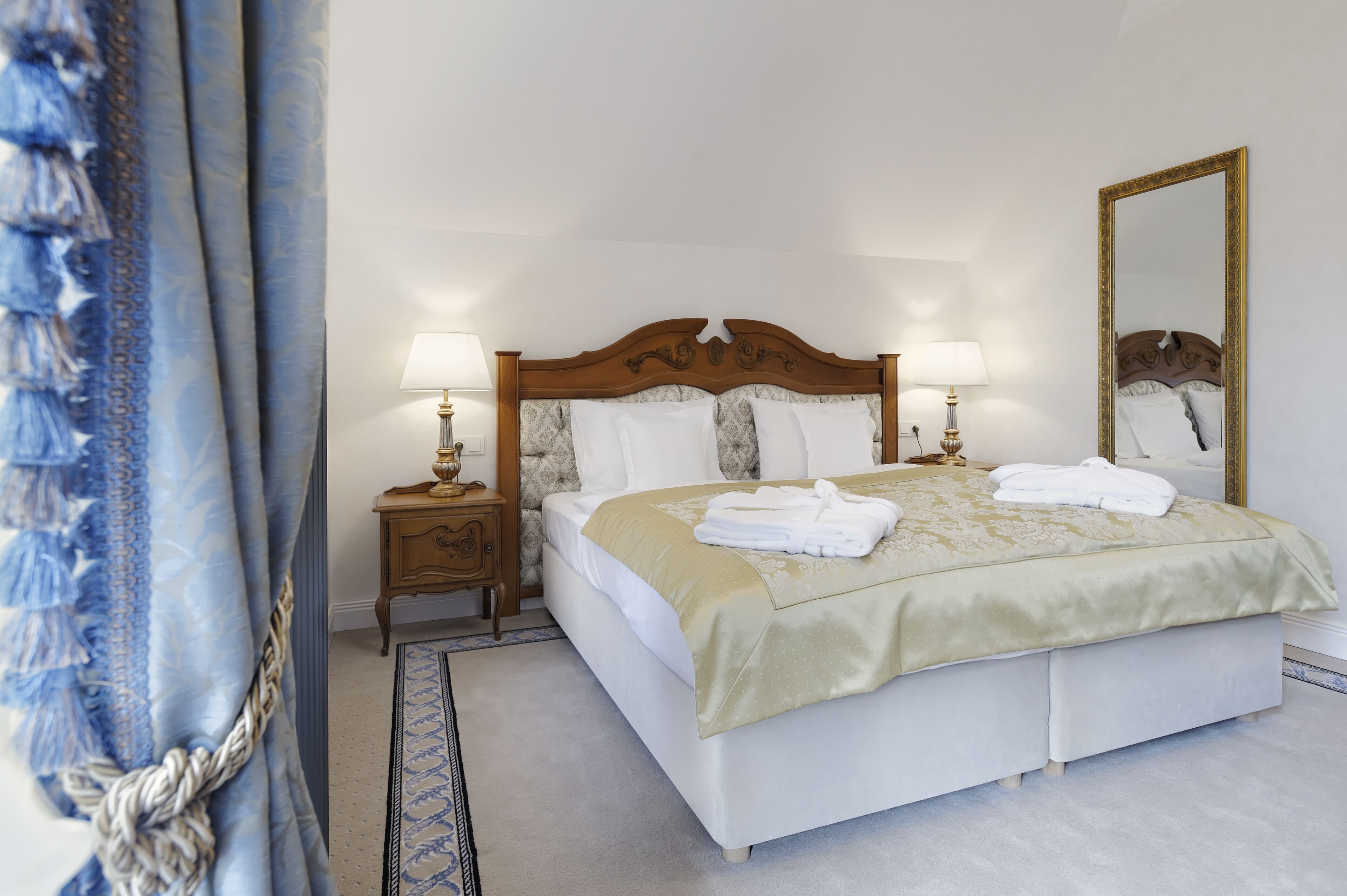 Un lit bien fait, avec du linge de lit blanc et une couverture beige, deux lampes de chevet, une table de chevet en bois et un grand miroir au mur.