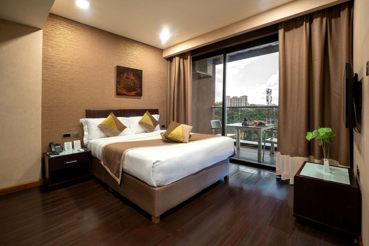 A modern hotel room with a large bed, a bedside table with a phone and a sign, a small table with a plant, and a view of the city through a large window.