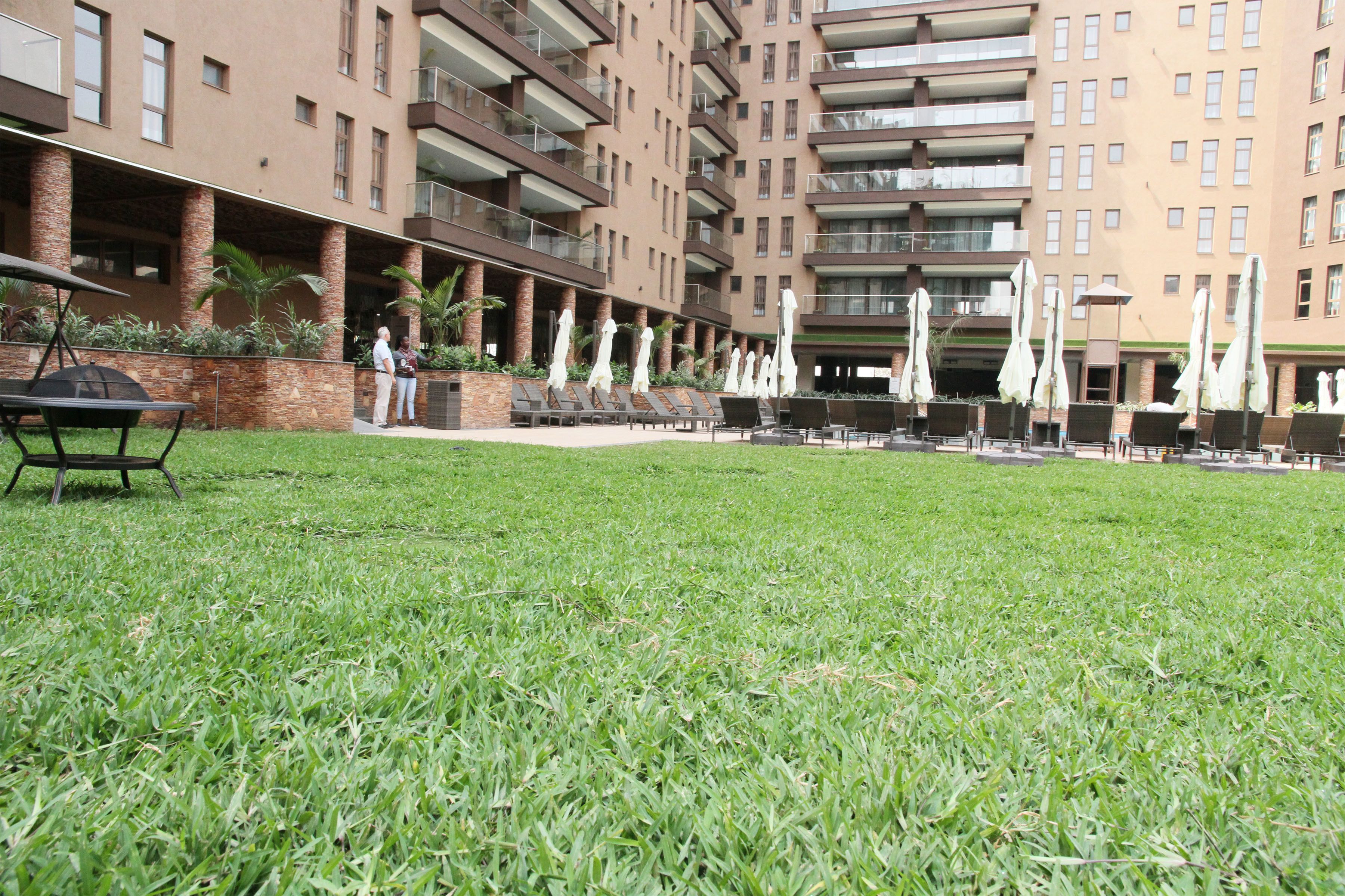 A grassy courtyard with outdoor seating and umbrellas, surrounded by a multi-story building with balconies.