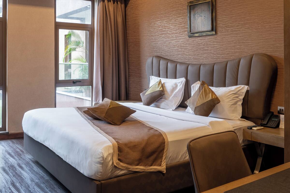 A hotel room with a large bed, a bedside table with a phone, and a view of palm trees through the window.