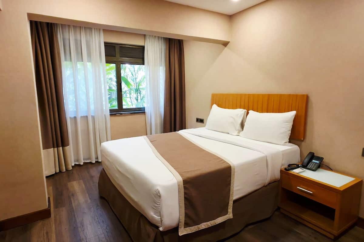 A hotel room with a bed, a bedside table with a phone, a TV mounted on the wall, and a window with curtains.
