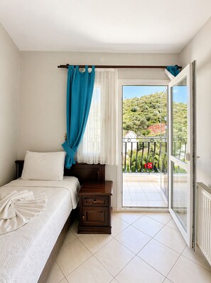 A bedroom with a bed, a nightstand, a window with curtains, and a balcony with a view of greenery and a flag.