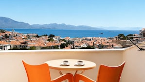 A balcony with a round table and two orange chairs, overlooking a coastal town with red-roofed buildings and a clear blue sea. - Villa Anfora Otel (Datça)