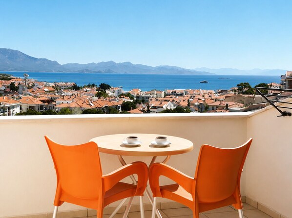 A balcony with a round table and two orange chairs, overlooking a coastal town with red-roofed buildings and a clear blue sea. - Villa Anfora Otel (Datça)