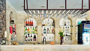 A bar area with two wicker chairs and a wooden stool, a shelf with various bottles, and a decorative wall with a stone-like texture. - Villa Anfora Otel (Datça)